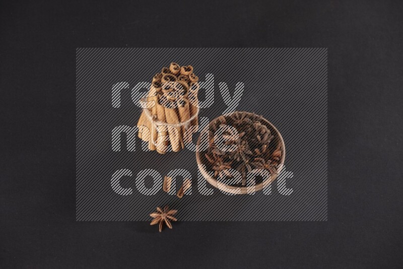 A stacked and bounded cinnamon sticks and a wooden bowl full of star anise on a black background