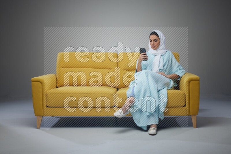 A Saudi woman wearing a light blue Abaya and white head scarf sitting on a yellow sofa and texting on her phone on a grey background