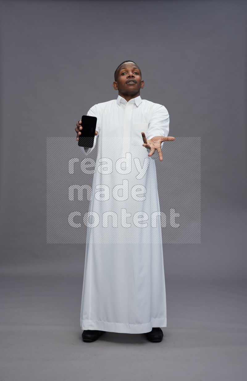 Saudi man wearing thob standing showing phone to camera on gray background