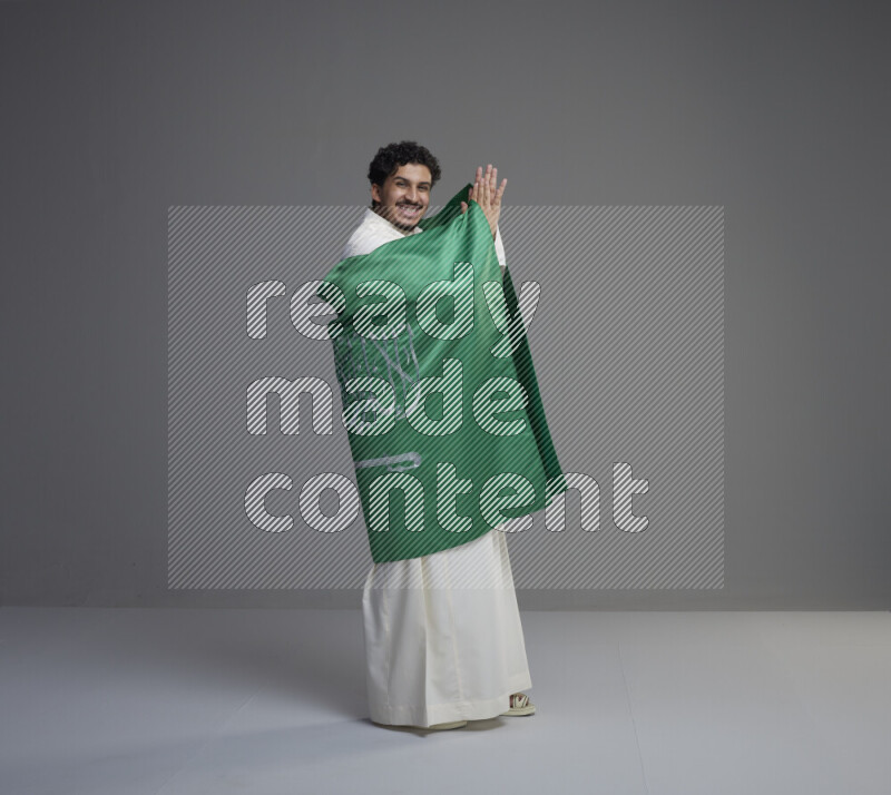 A Saudi man standing wearing thob wrapping big Saudi flag on gray background