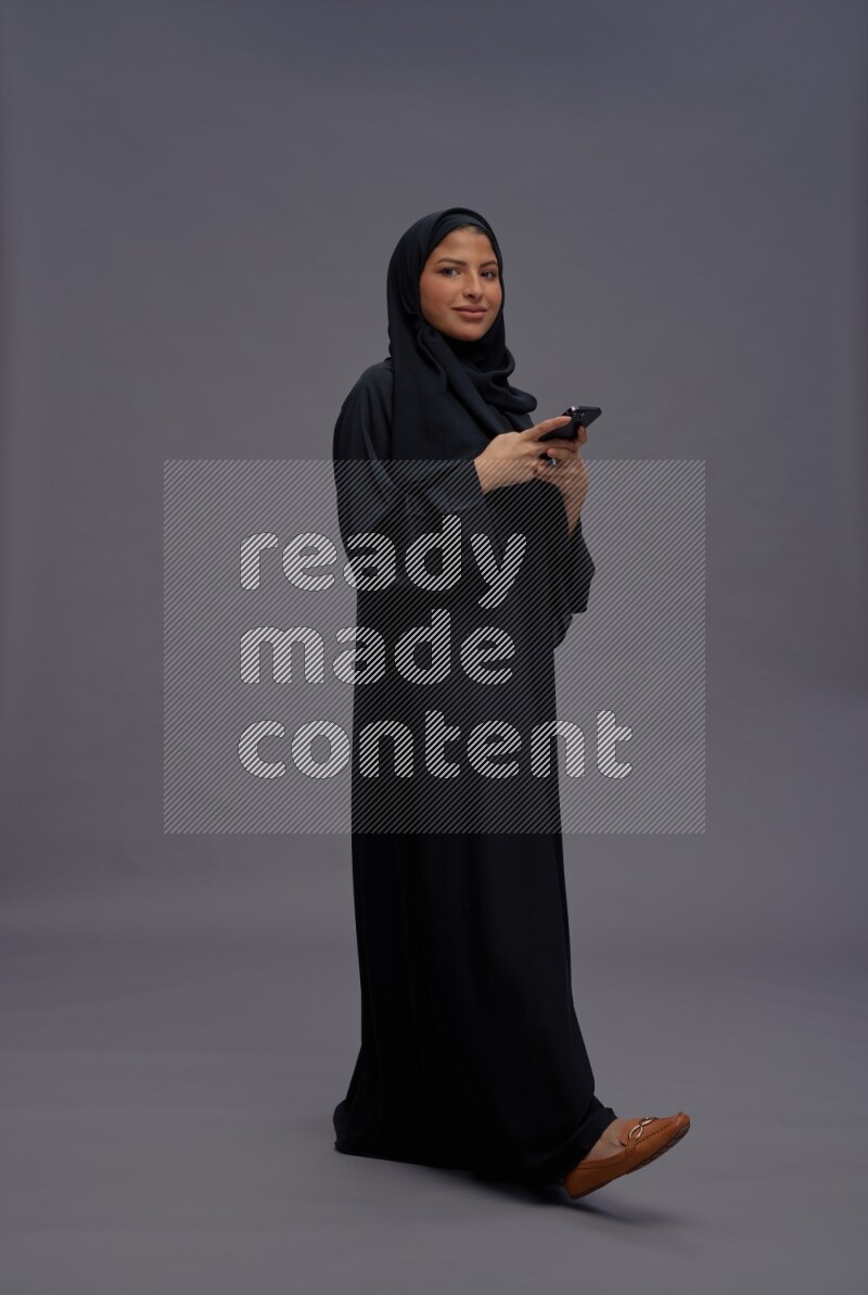 Saudi woman wearing Abaya standing texting on phone on gray background
