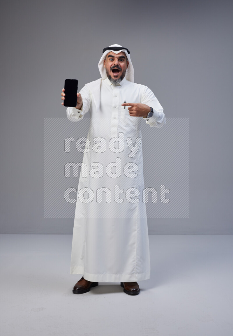 Saudi man Wearing Thob and white Shomag standing showing phone to camera on Gray background