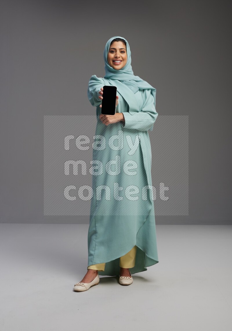 Saudi Woman wearing Abaya standing showing phone to camera on Gray background