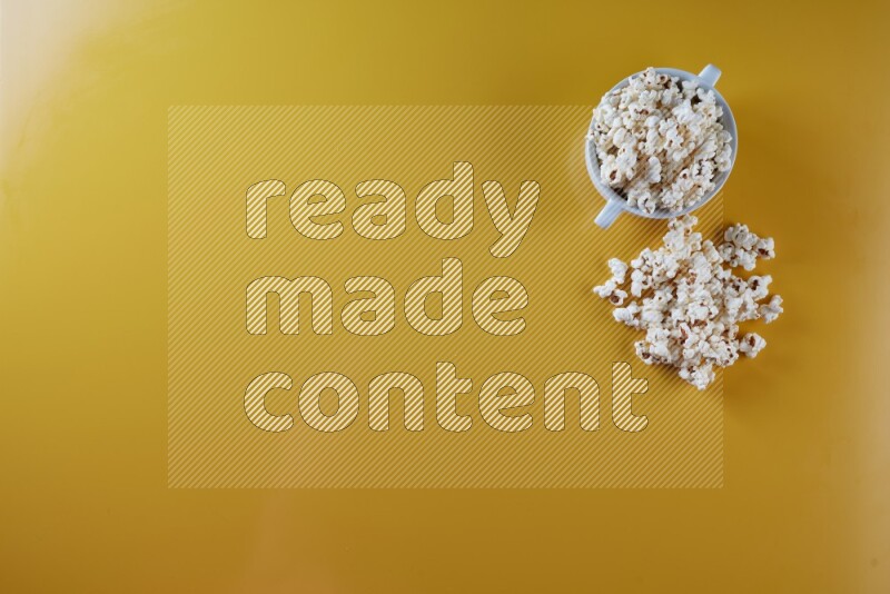 A white ceramic bowl full of popcorn with popcorn beside it on a yellow background in a top view shot