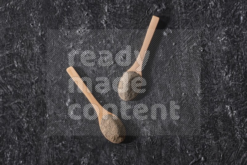 2 wooden spoons full of black pepper powder on a textured black flooring