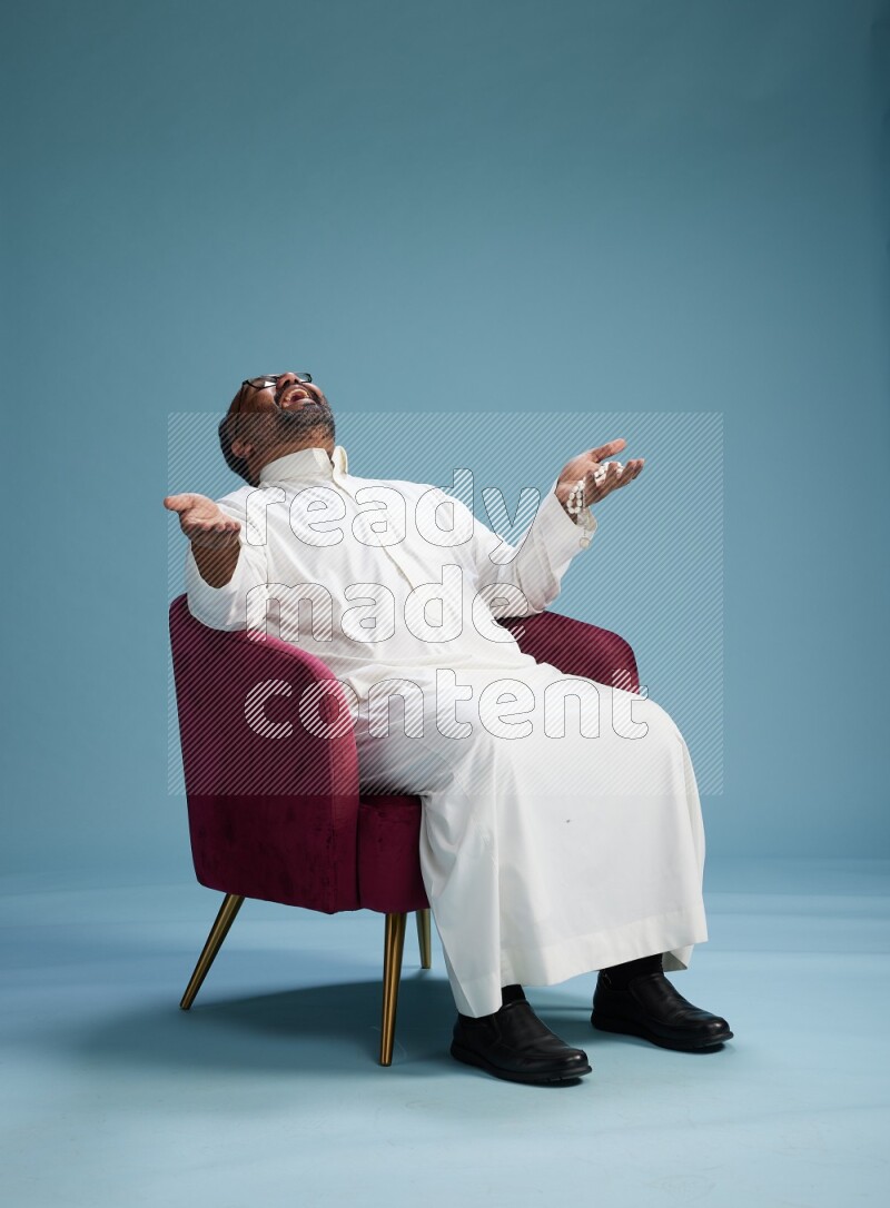 Saudi Man without shimag sitting on chair Interacting with the camera on blue background