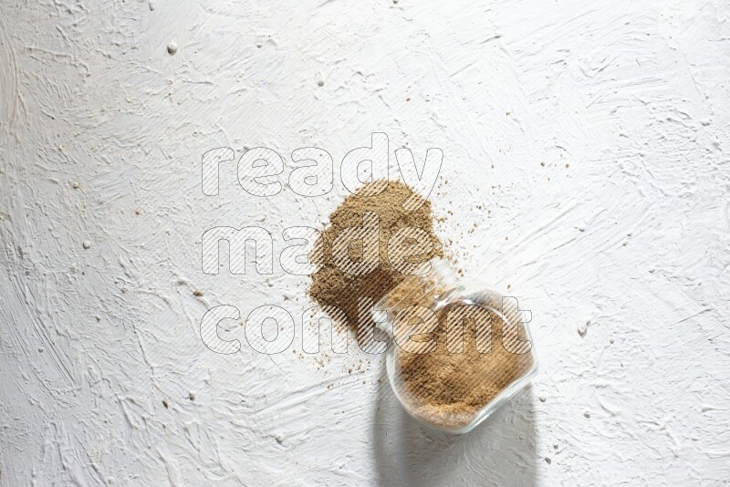 وعاء زجاجي مقلوب ممتلئ ببودرة الكمون والبودرة تناثرت منه على خلفية بيضاء