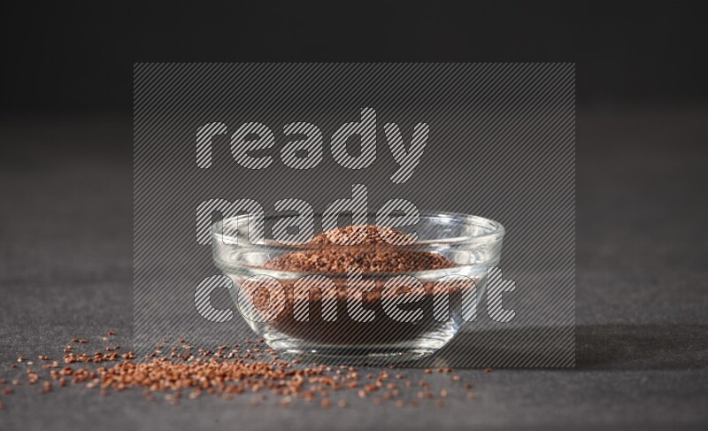 A glass bowl full of garden cress seeds surrounded by seeds on a black flooring