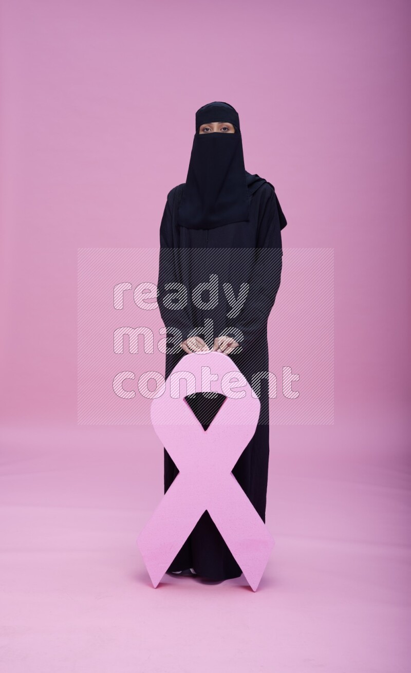 Saudi woman wearing abaya and niqab standing holding awareness ribbon on pink background