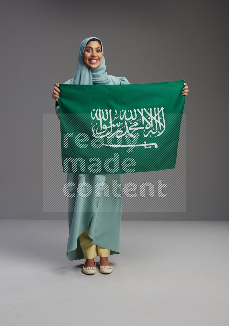 Saudi Woman wearing Abaya standing holding Saudi flag on Gray background