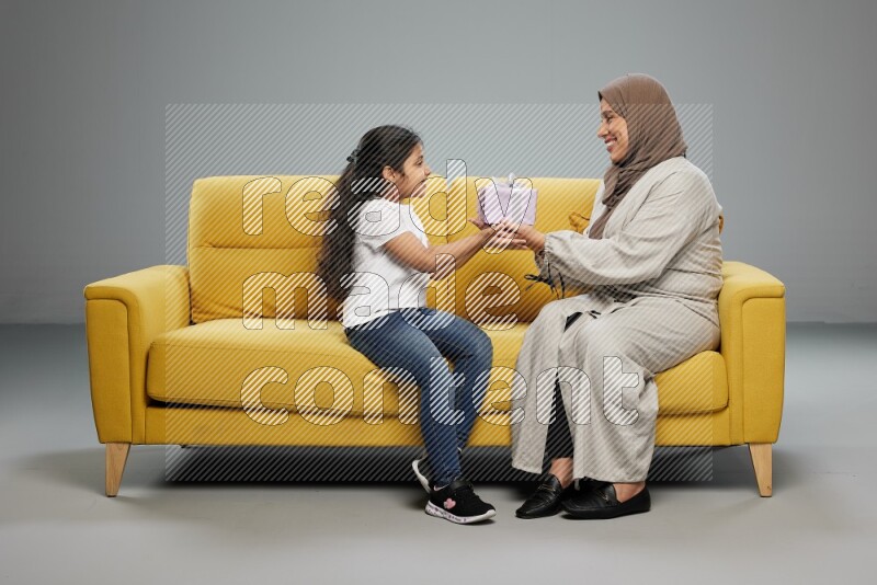 A girl sitting on a yellow sofa and giving a gift to her mother on gray background