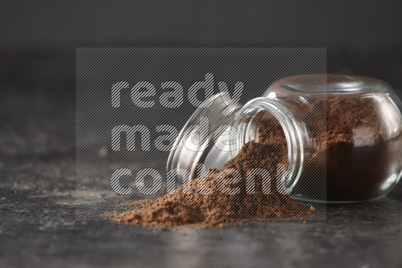 A flipped glass spice jar full of cloves powder and powder came out of it on textured black flooring