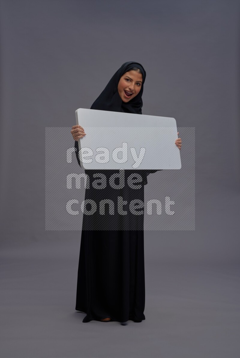 Saudi woman wearing Abaya standing holding white board on gray background