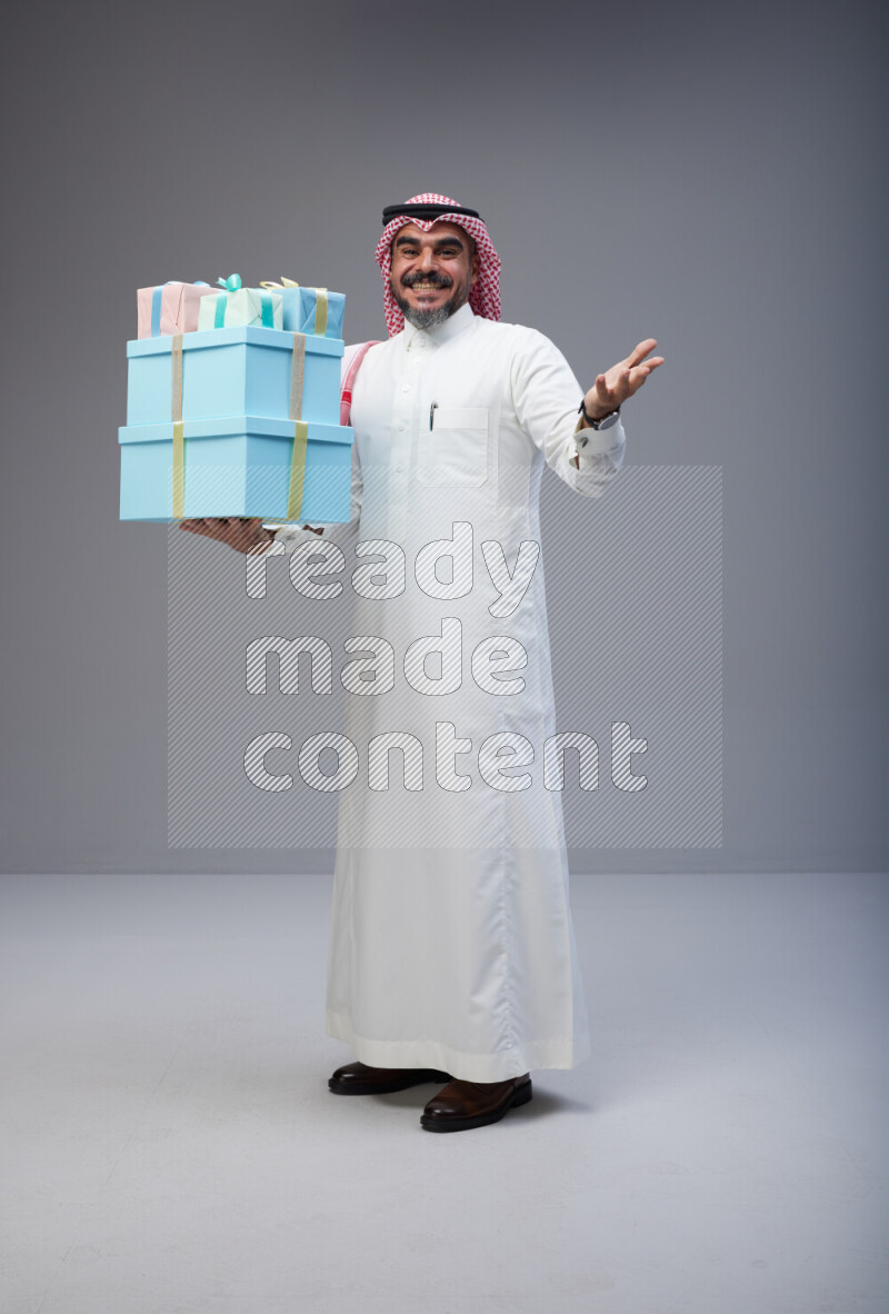 Saudi man Wearing Thob and red Shomag standing holding gift box on Gray background