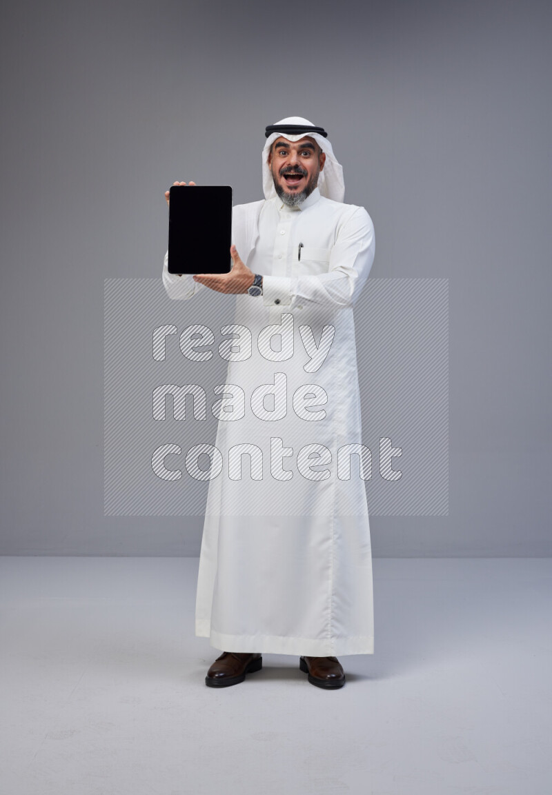 Saudi man Wearing Thob and white Shomag standing showing tablet to camera on Gray background