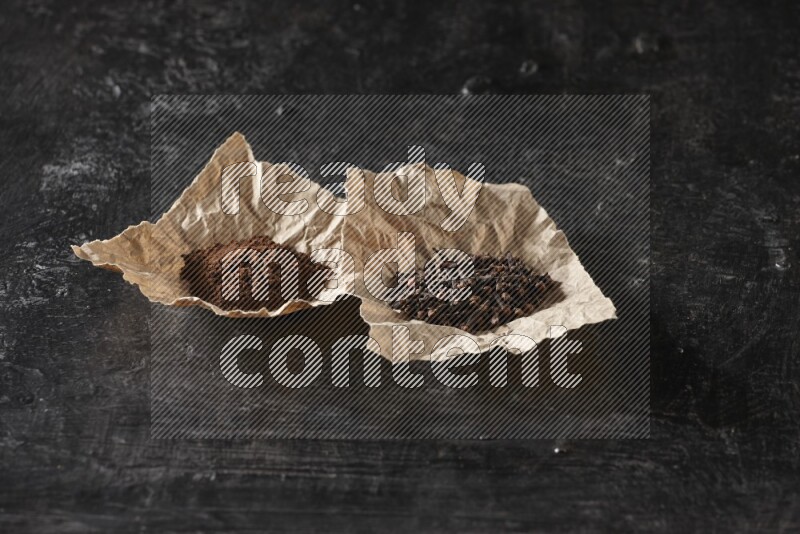 Cloves powder and cloves in 2 crumpled pieces of paper on a textured black flooring