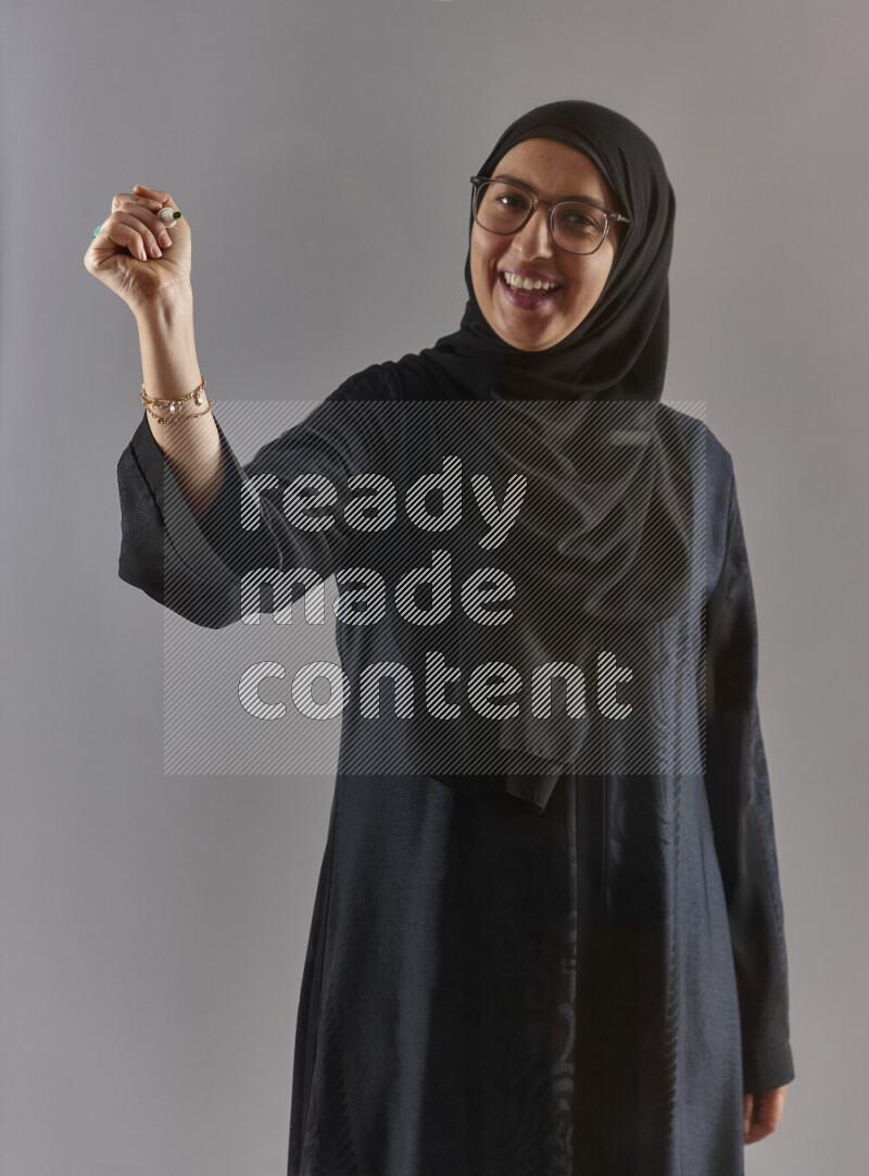Woman in black abaya, hijab and glasses holding a marker pen to write on a transparent board with different reactions and expressions on grey background