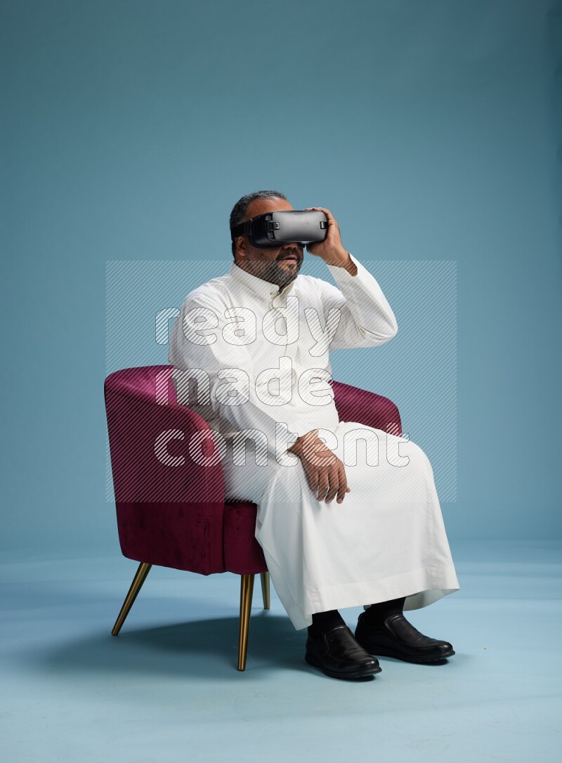Saudi Man without shimag sitting on chair  gaming with VR on blue background