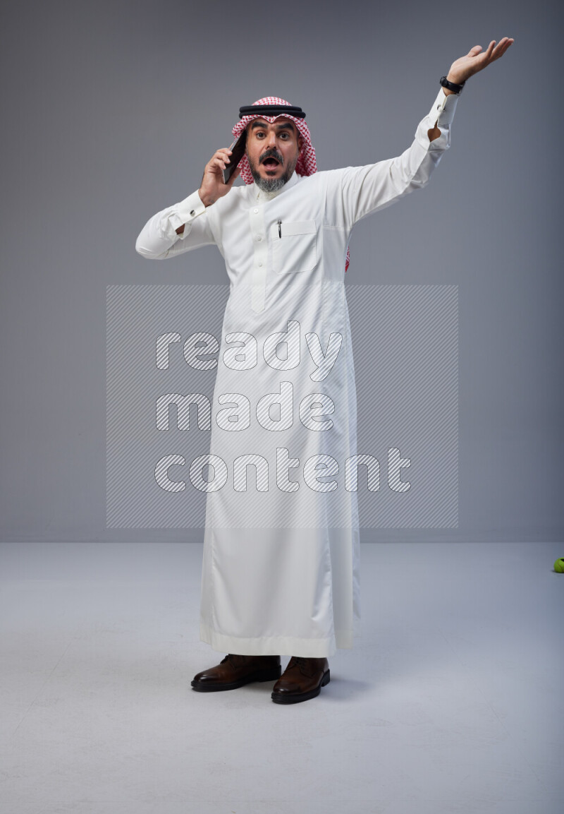 Saudi man Wearing Thob and red Shomag standing talking on phone on Gray background