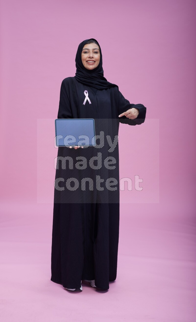 Saudi woman wearing pink ribbon on Abaya standing showing a tablet to camera on pink background