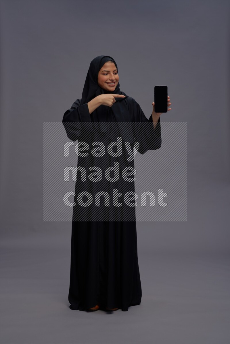 Saudi woman wearing Abaya standing showing phone to camera on gray background