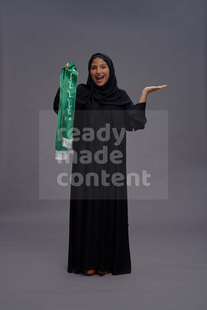 Saudi woman wearing Abaya standing holding Saudi flag on gray background