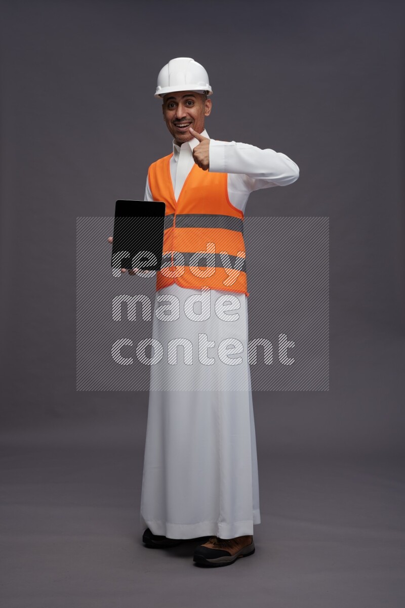 Saudi man wearing thob with engineer vest standing showing tablet to camera on gray background