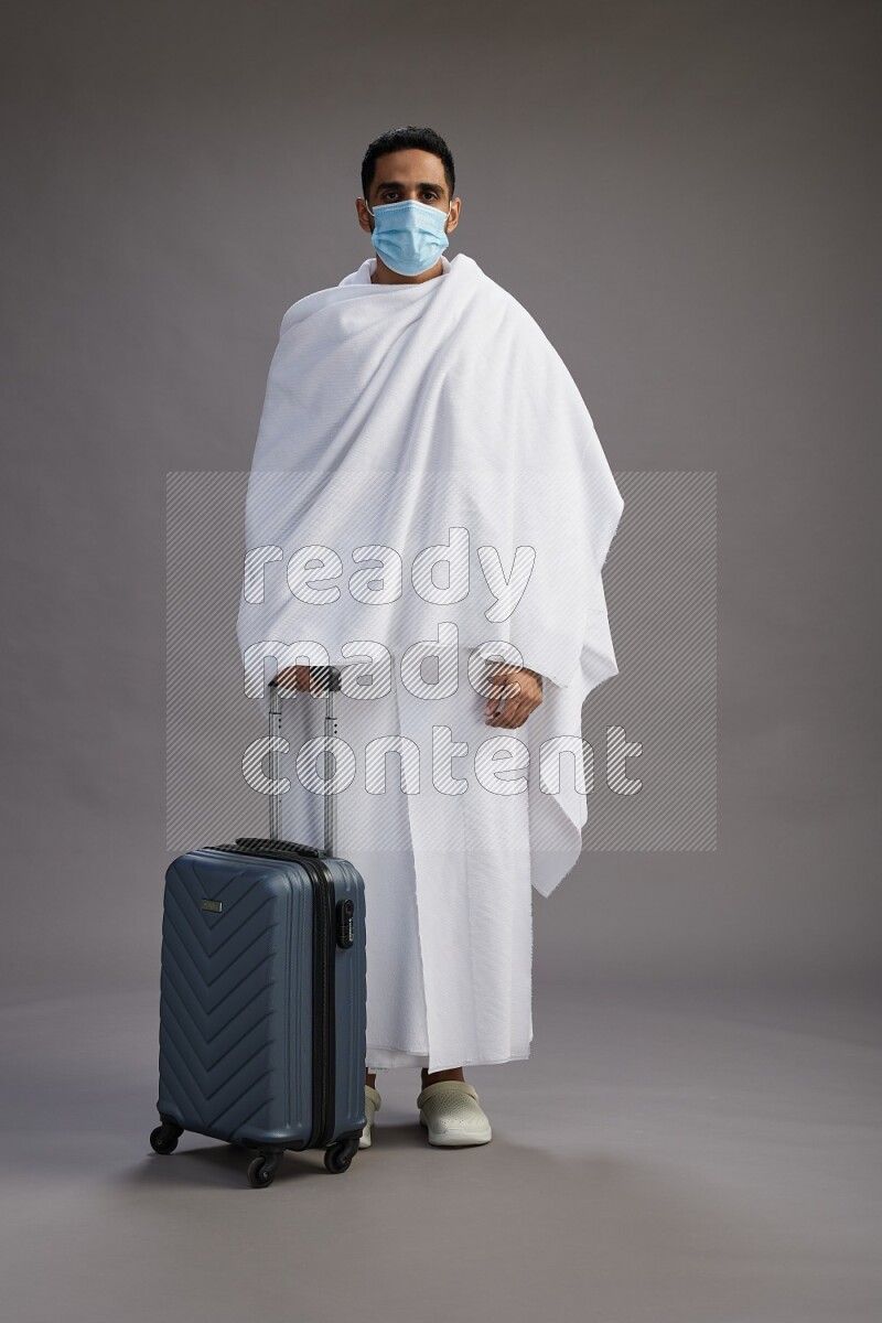 A man wearing Ehram with face mask Standing holding traveling bag on gray background