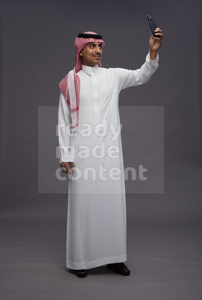 Saudi man wearing thob and shomag standing taking selfie on gray background