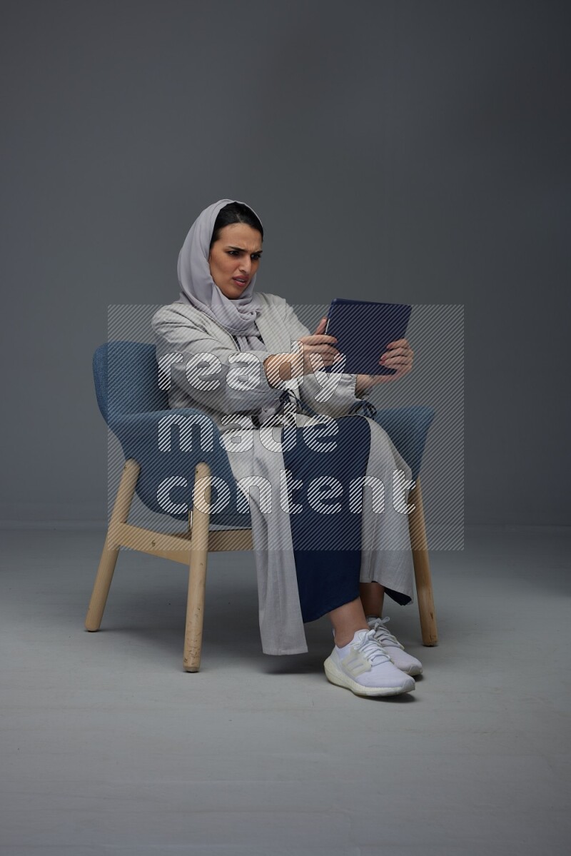 A Saudi female wearing a light gray Abaya and head scarf sitting on a dark grey chair while holding her iPad on a grey background