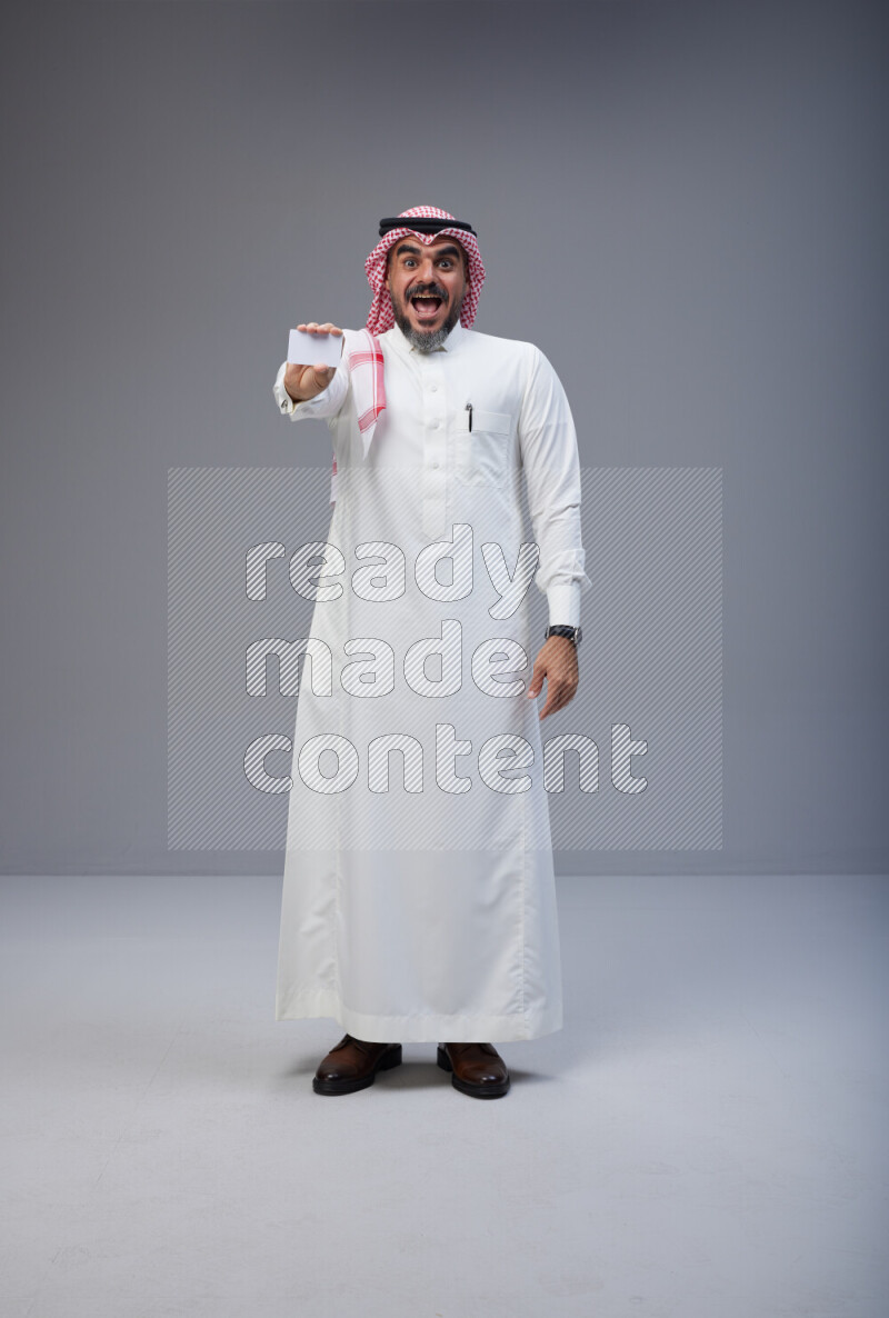 Saudi man Wearing Thob and red Shomag standing holding ATM card on Gray background