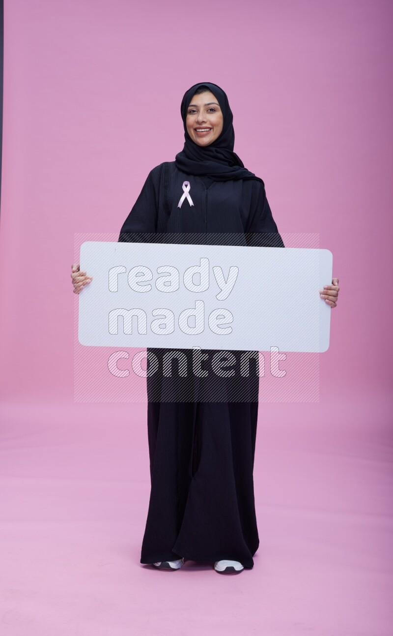 Saudi woman wearing pink ribbon on Abaya standing holding board on pink background