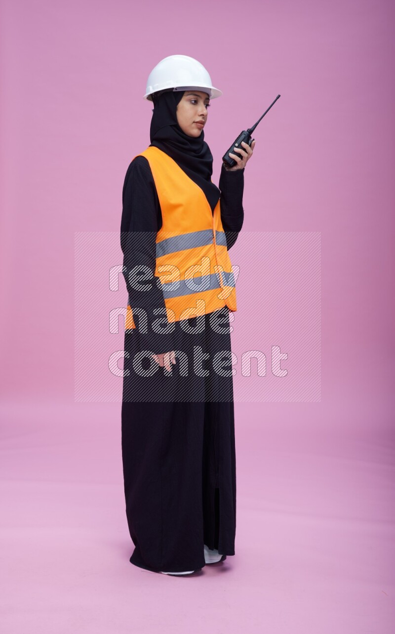 Saudi woman wearing Abaya with engineer vest and helmet standing holding walkie-talkie on pink background