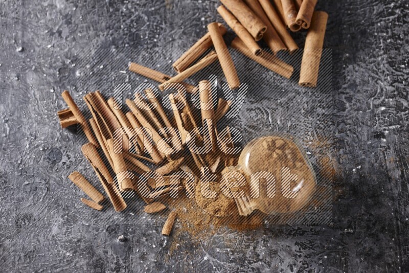 Herbal glass jar full cinnamon powder flipped and a metal spoon full of powder surrounded by cinnamon sticks on textured black background in different angles