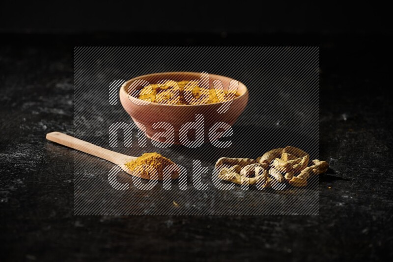 A wooden bowl and a wooden spoon full of turmeric powder with dried turmeric fingers on textured black flooring