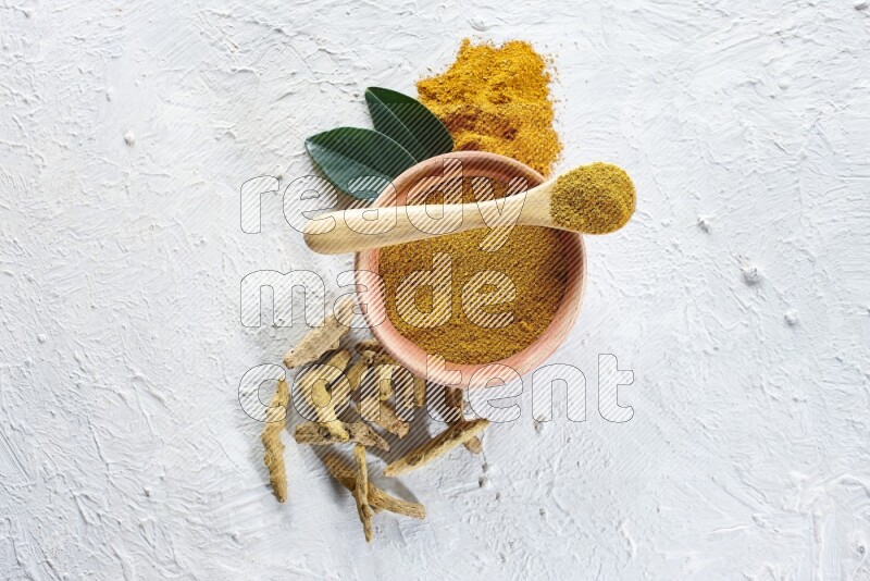 A wooden bowl and wooden spoon full of turmeric powder with dried turmeric fingers beside it on textured white flooring