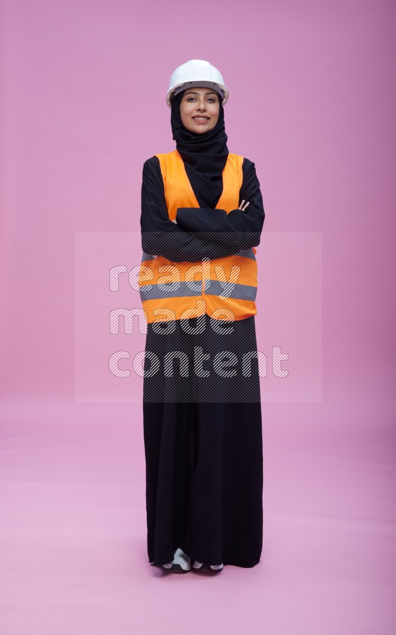 Saudi woman wearing Abaya with engineer vest and helmet standing with crossed arms on pink background