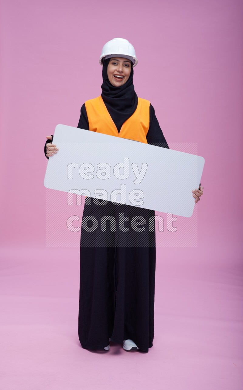 Saudi woman wearing Abaya with engineer vest and helmet standing holding board on pink background
