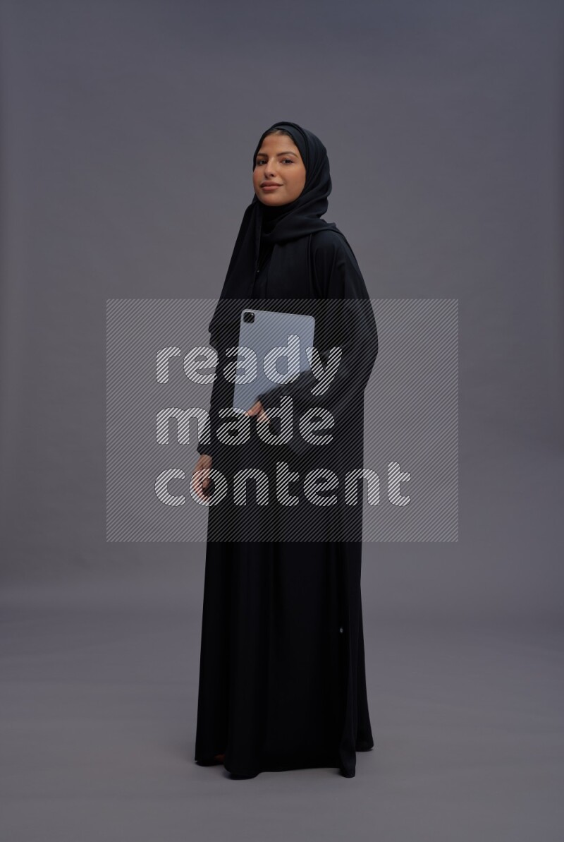 Saudi woman wearing Abaya standing working on tablet on gray background