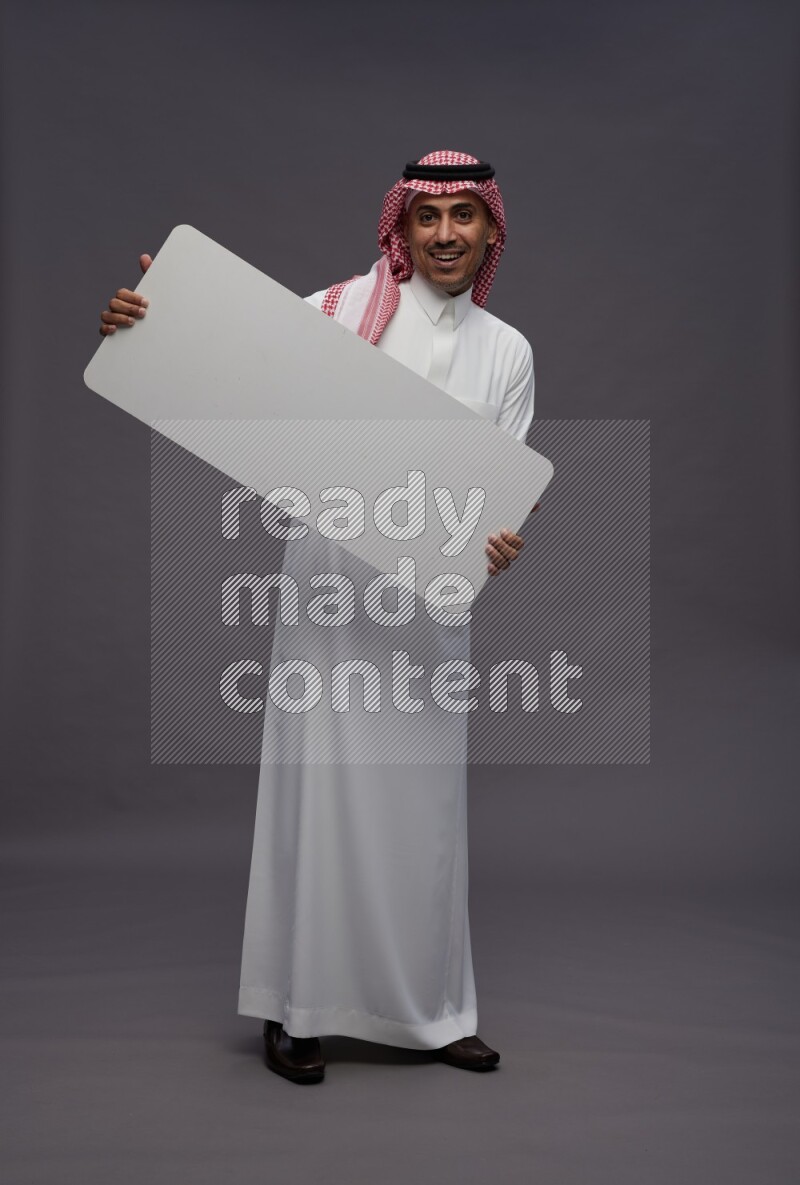 Saudi man wearing thob and shomag standing holding board on gray background
