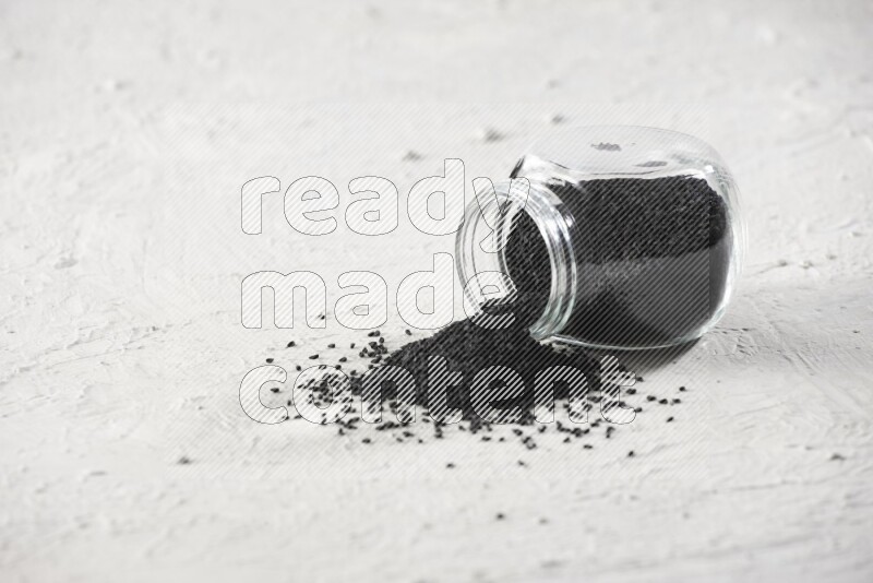 A glass spice jar full of black seeds and the jar flipped and seeds spread on a textured white flooring