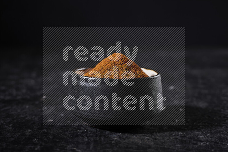 A black pottery bowl full of ground paprika powder on black background