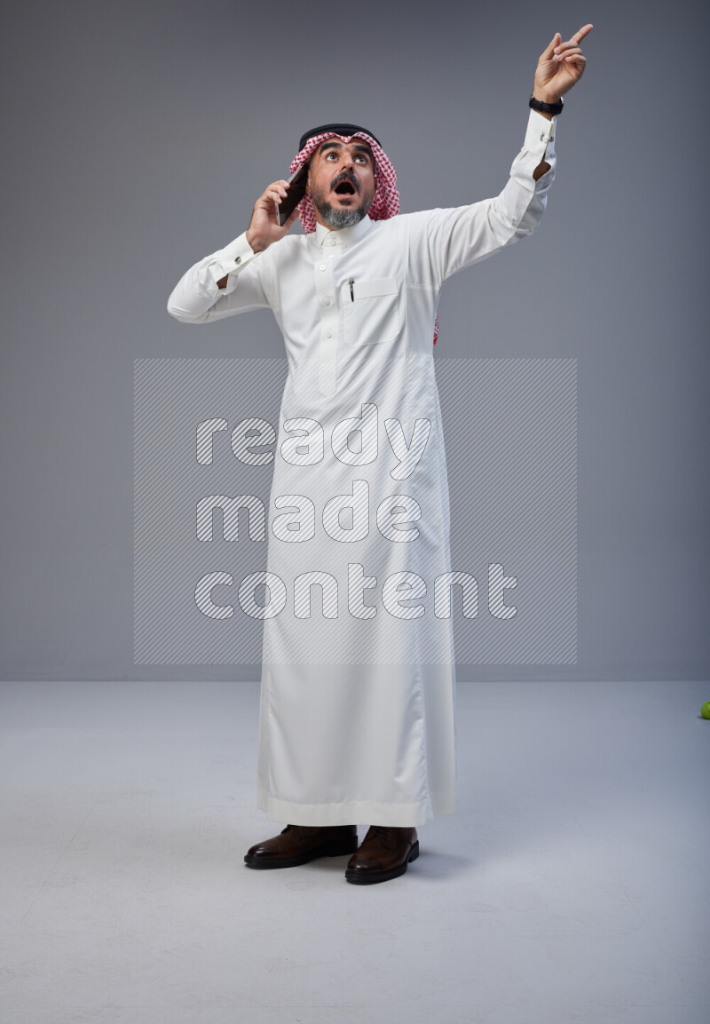 Saudi man Wearing Thob and red Shomag standing talking on phone on Gray background