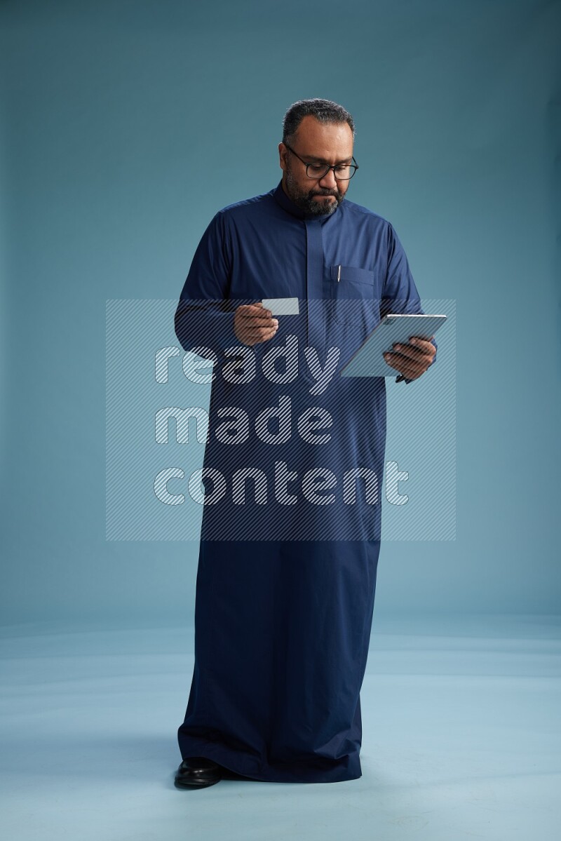 Saudi Man without shimag Standing holding ATM card while working on tablet on blue background
