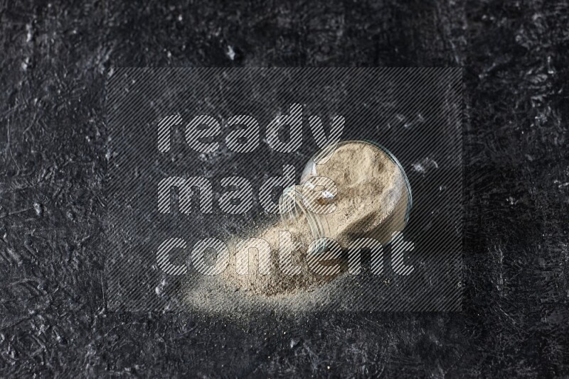 A flipped herbal glass jar full of white pepper powder with spilled powder on textured black flooring