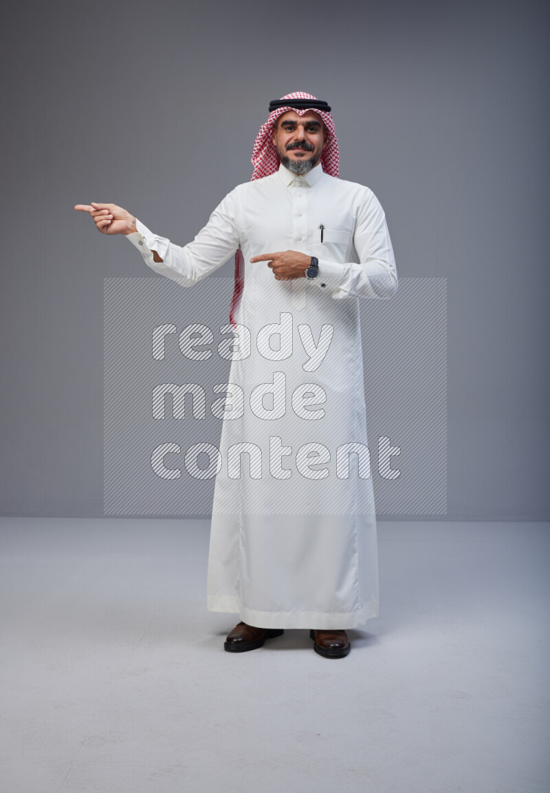 Saudi man Wearing Thob and red white Shomag standing interacting with the camera on Gray background