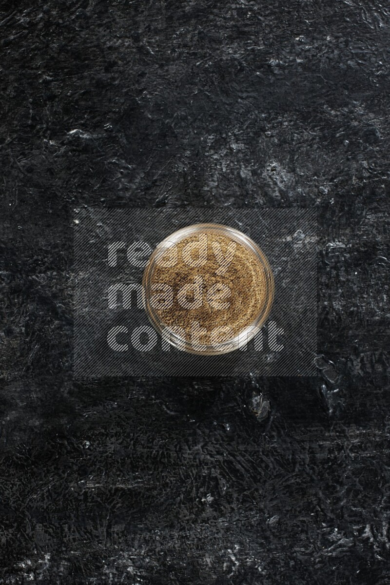 A glass jar full of cumin powder on a textured black flooring