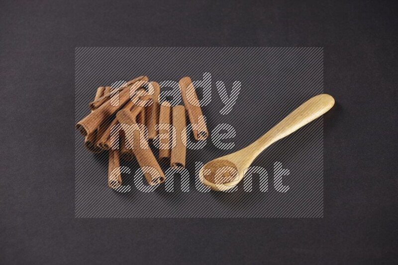 Cinnamon sticks stacked beside a wooden spoon full of cinnamon powder on black background