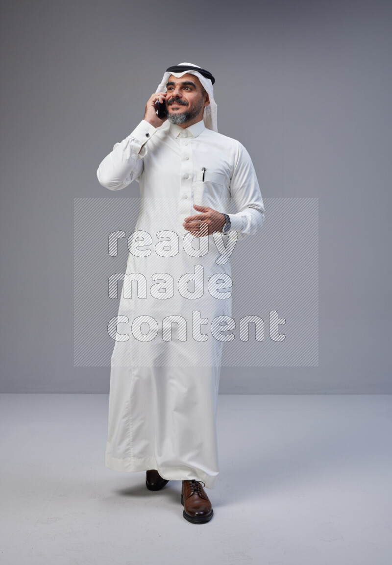 Saudi man Wearing Thob and white Shomag standing talking on phone on Gray background