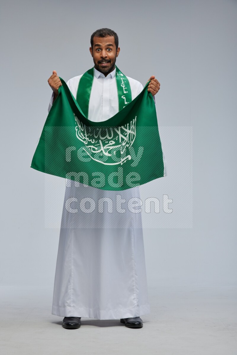 Saudi man wearing Thob standing holding Saudi flag on Gray background